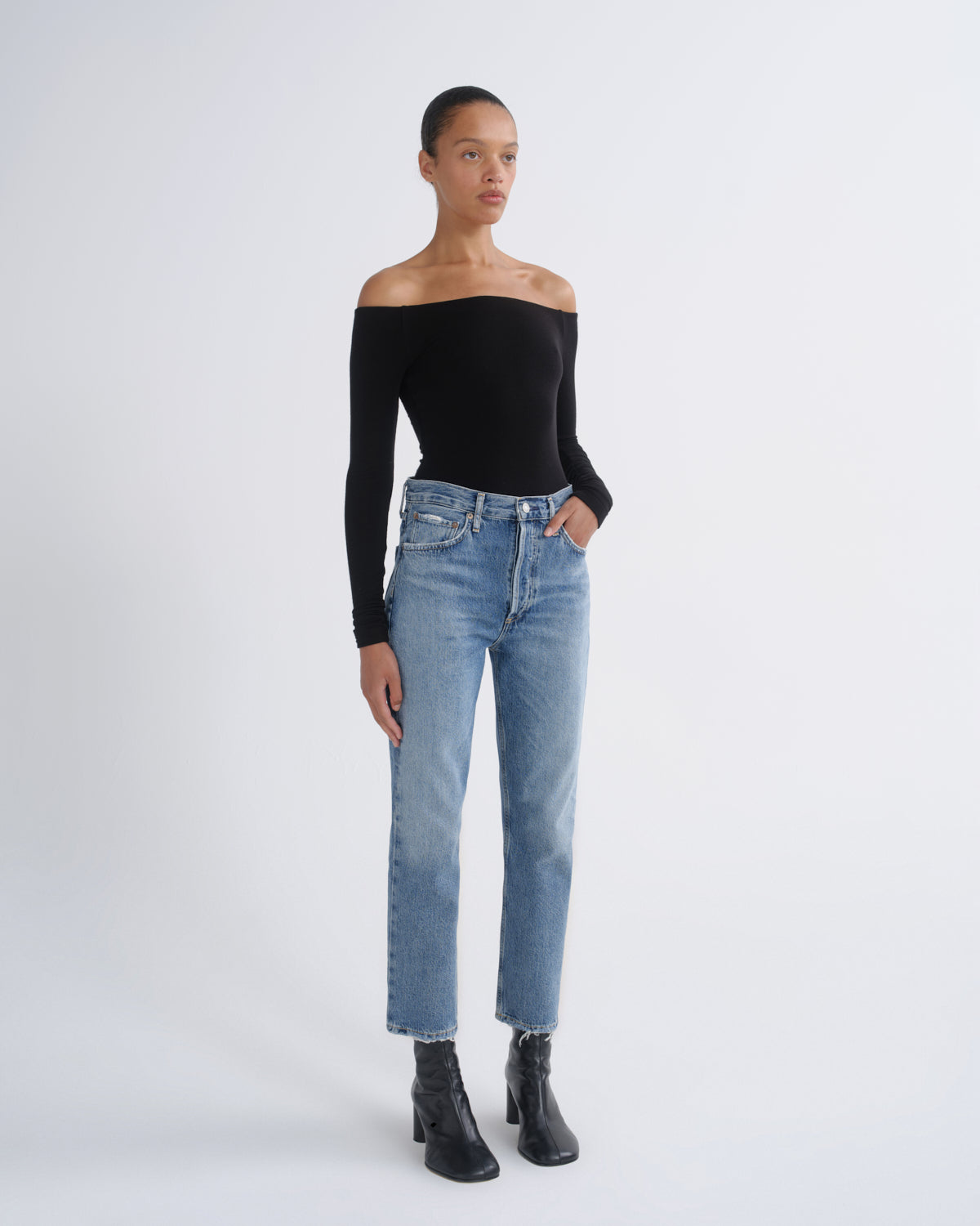 A model stands against a light gray studio background wearing a black off-the-shoulder long-sleeve top, light-wash blue straight-leg jeans, and black ankle boots.
