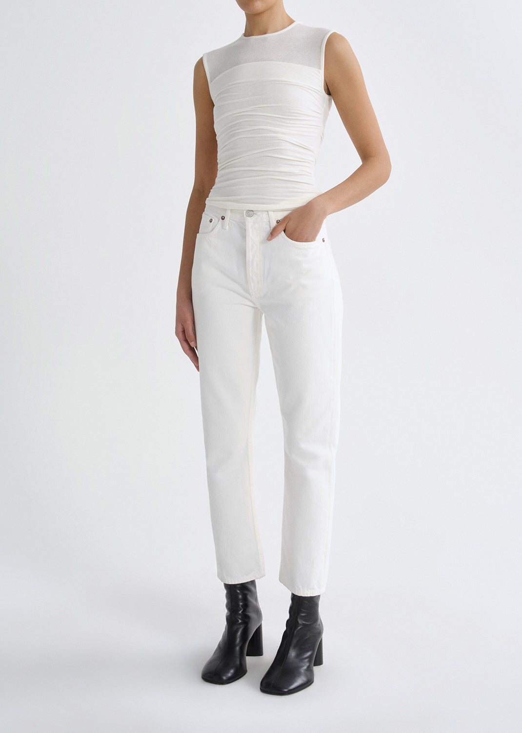 Person wearing a white sleeveless ruched top with white straight leg jeans and black ankle boots on a grey background