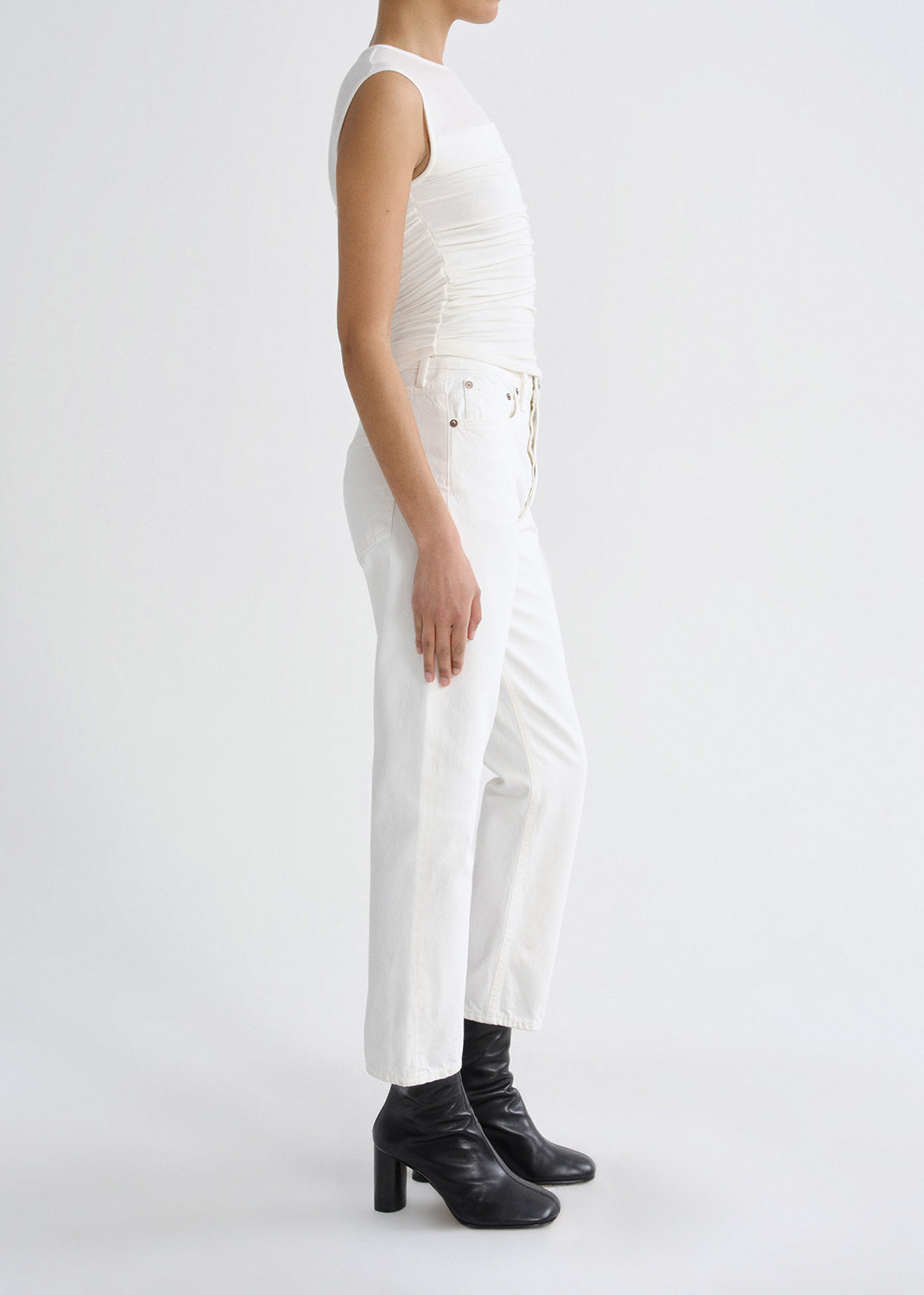 Person wearing a white sleeveless ruched top with white straight leg jeans viewed from the side on a grey background