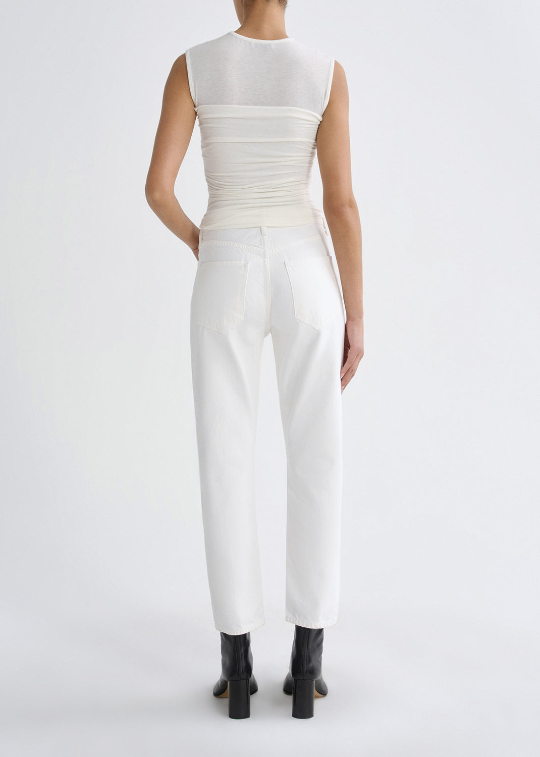 Person wearing a white sleeveless ruched top with white straight leg jeans viewed from the back on a grey background