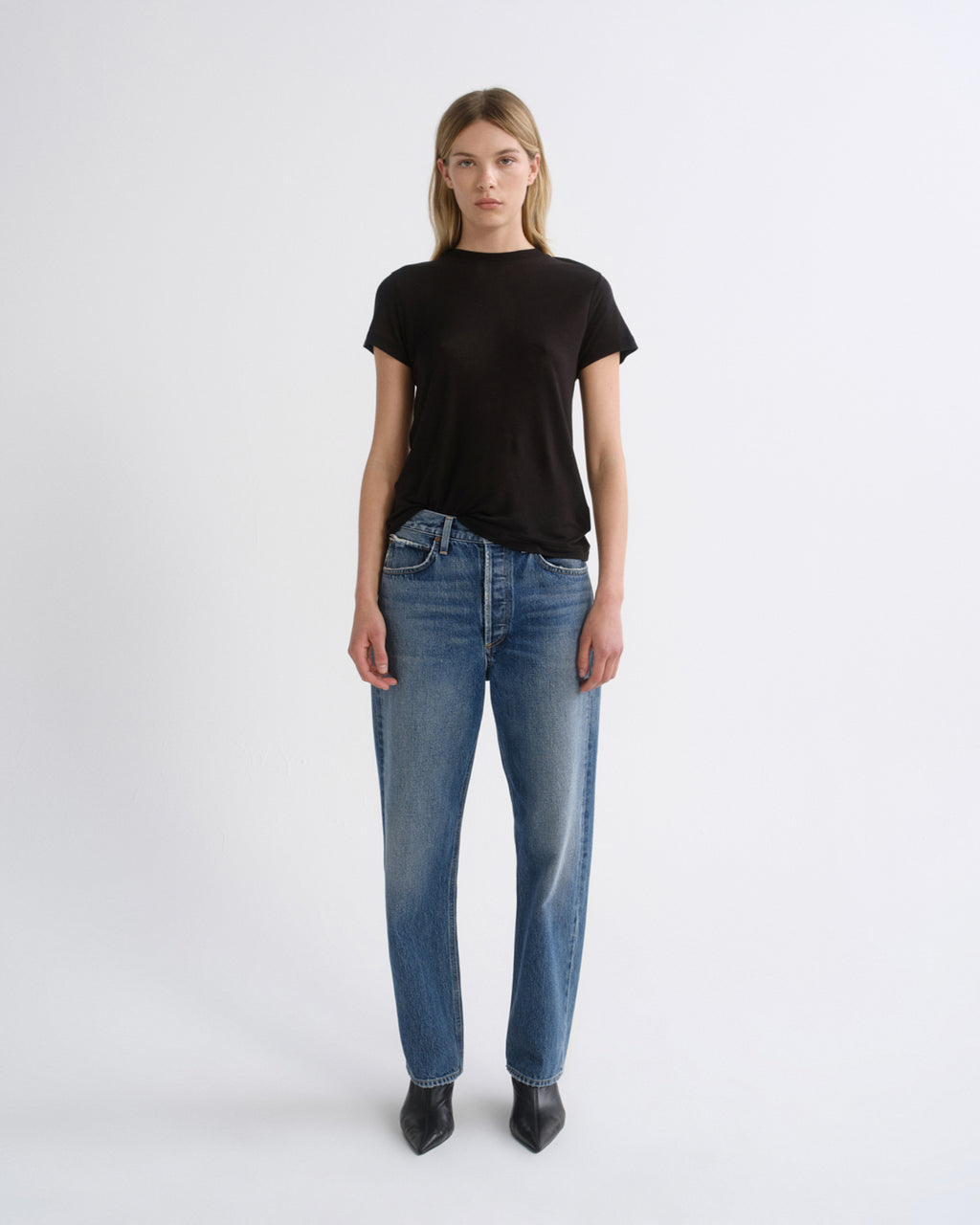A model stands against a light gray studio background wearing a black short-sleeve crewneck t-shirt, blue straight-leg jeans, and black pointed-toe ankle boots.
