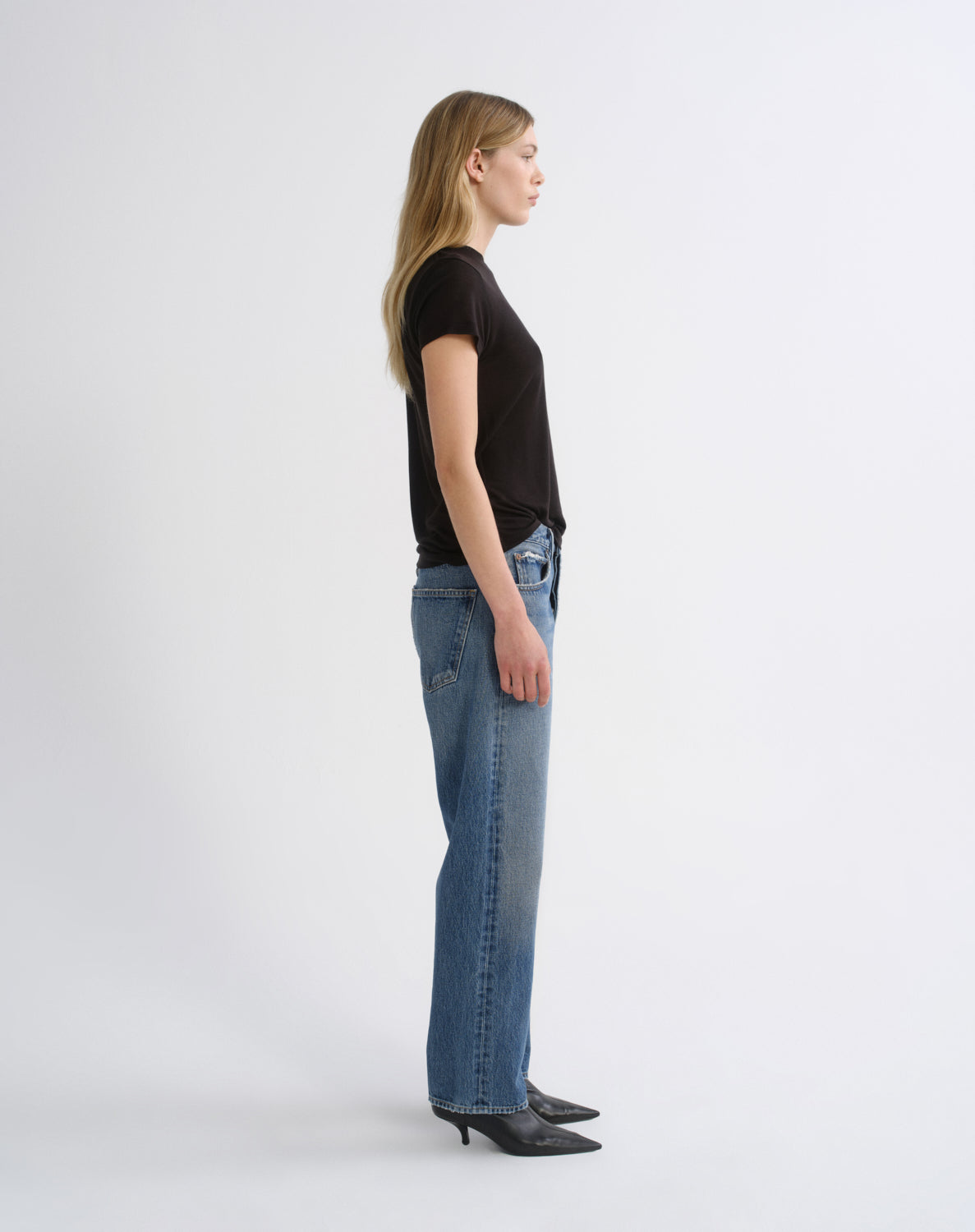 A woman in a black short-sleeve t-shirt, blue wide-leg jeans, and black pointed-toe heels, shown in a side-profile studio pose against a light gray background.
