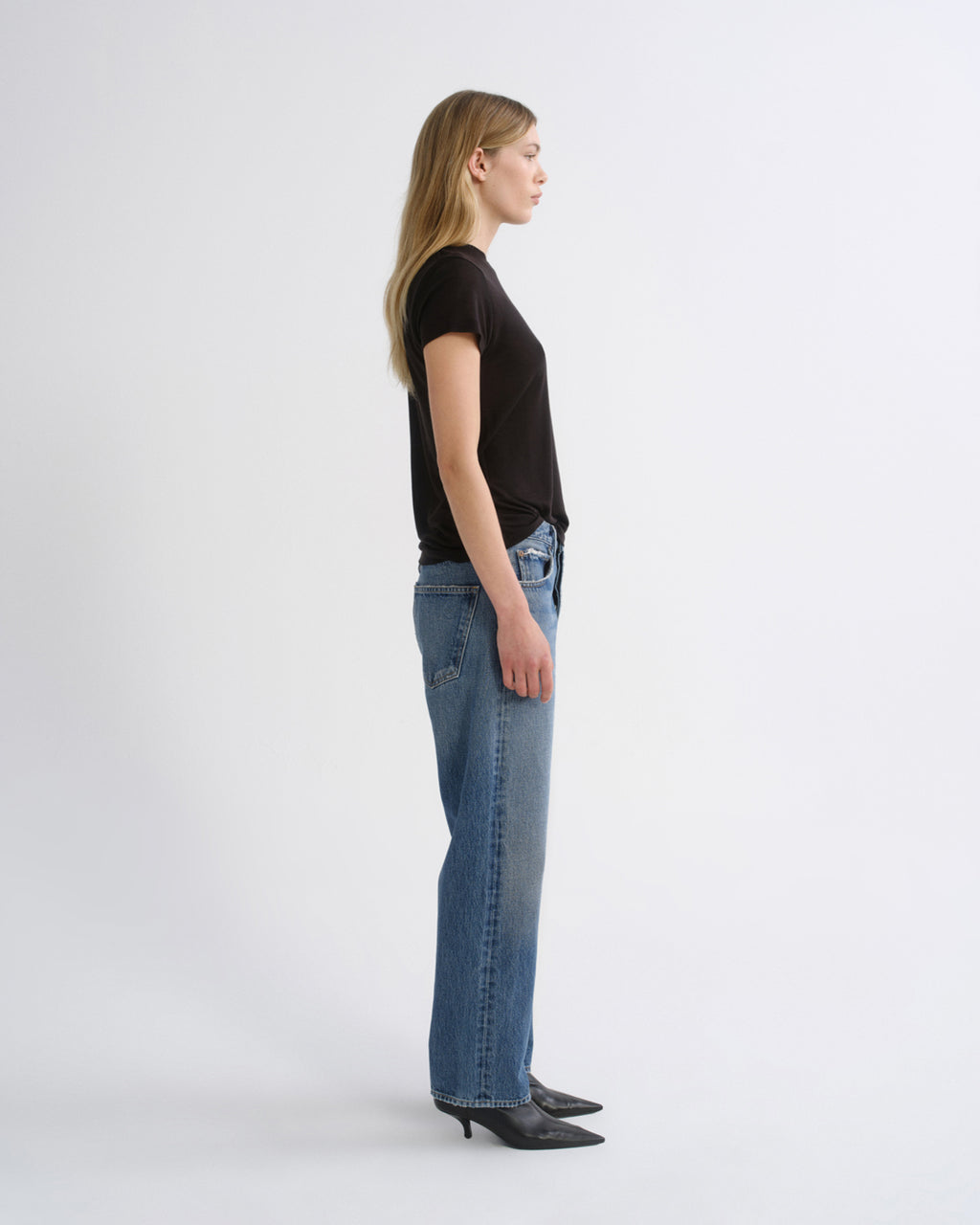 A woman standing in profile wearing a black short-sleeve t-shirt, blue straight-leg jeans, and black pointed-toe heels against a plain light gray studio background.