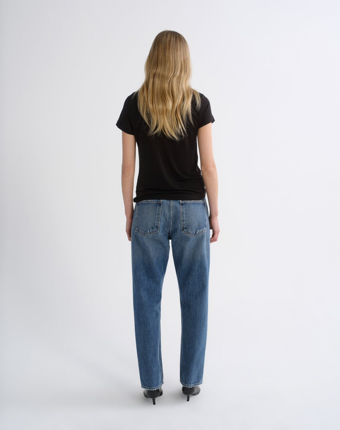 A woman viewed from behind wearing a black short-sleeve t-shirt and blue straight-leg jeans, standing against a plain light gray studio background.