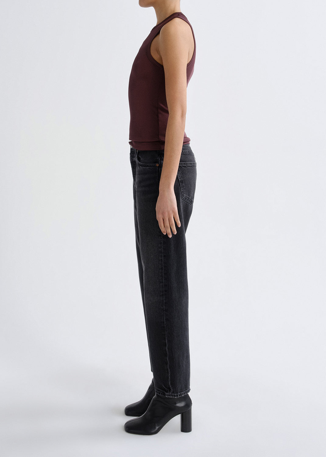 A side-profile studio shot of a model wearing a sleeveless dark burgundy top, black wide-leg jeans, and black heeled ankle boots against a grey background.