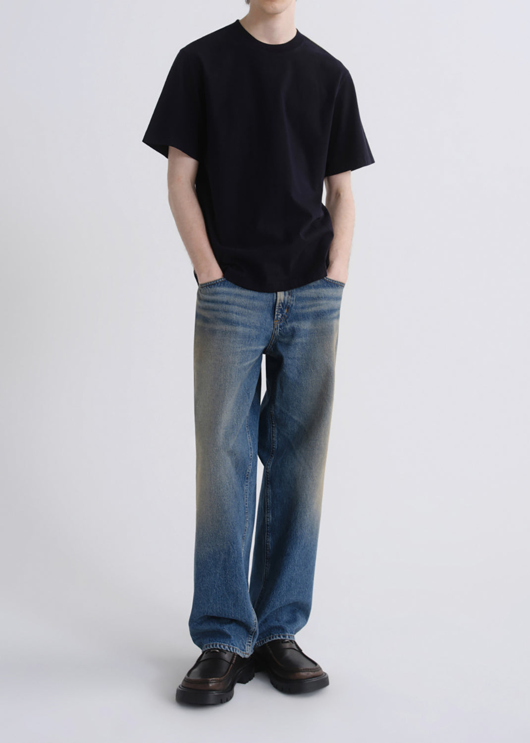 A model wearing a black short-sleeve crewneck t-shirt, blue wide-leg jeans with a faded wash, and dark brown leather loafers, standing against a grey background.