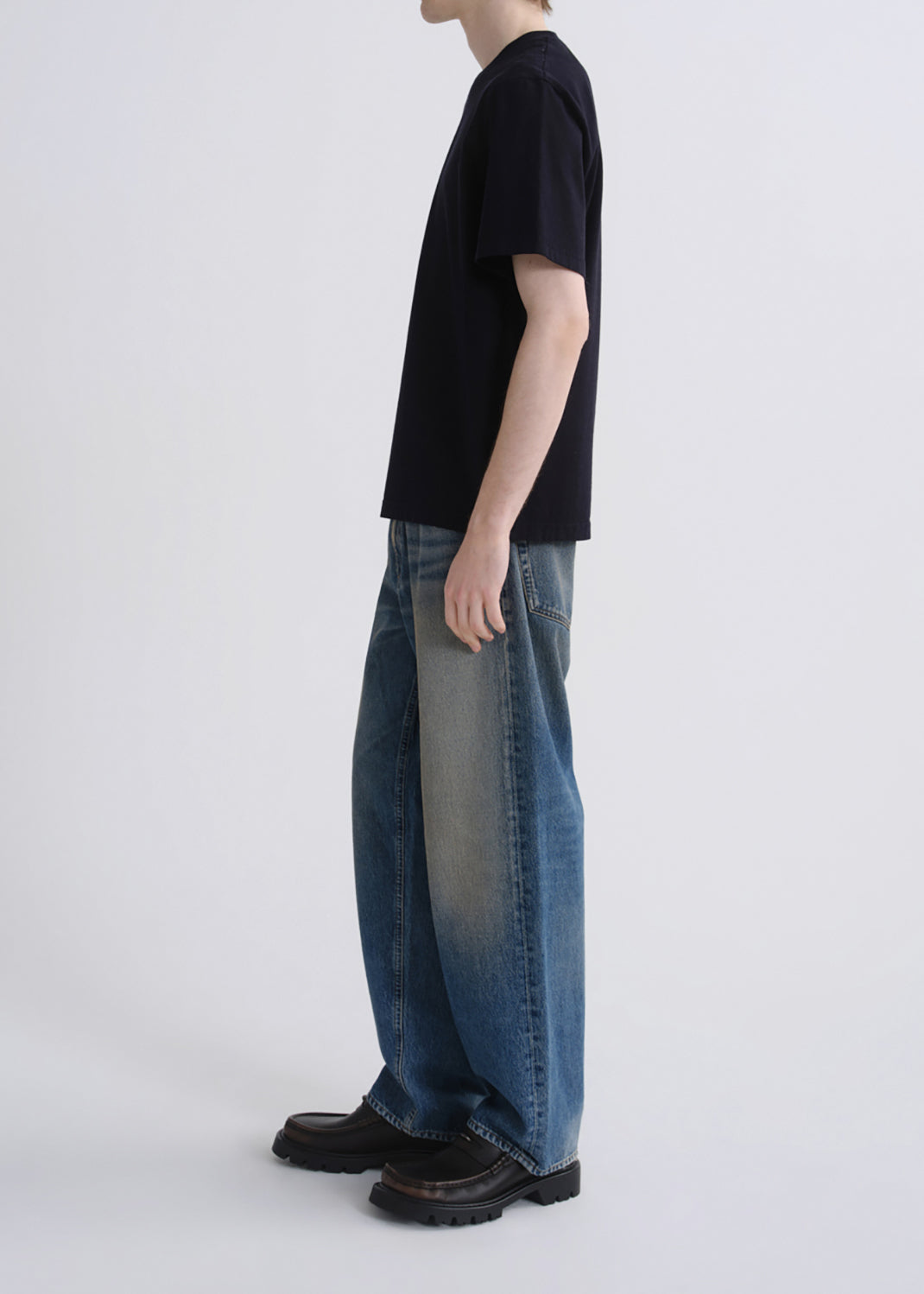 A side-profile studio shot of a model wearing a black short-sleeve t-shirt, wide-leg faded blue denim jeans, and black leather boots against a grey background.