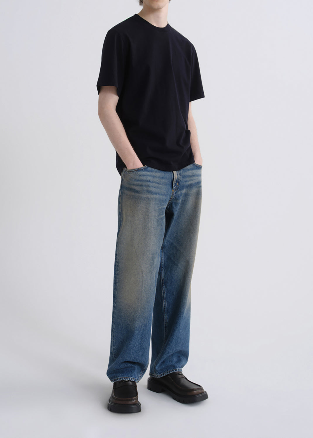 A model wearing a black short-sleeve crewneck t-shirt, blue wide-leg faded jeans, and black leather shoes, standing against a grey background.