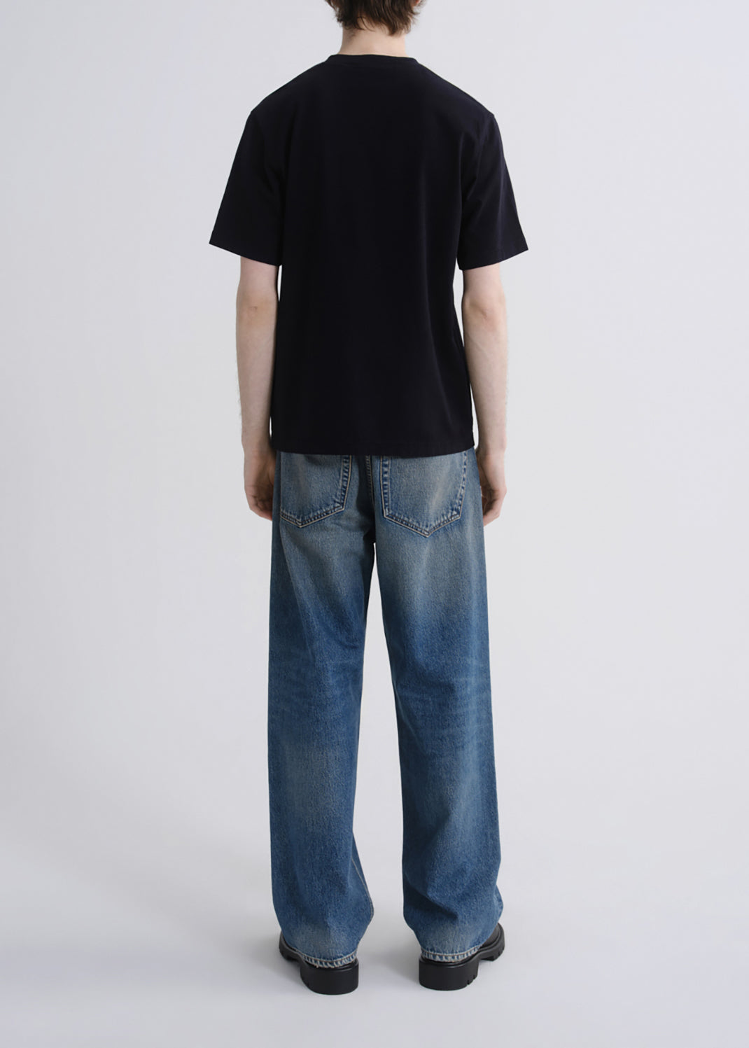 A person shown from the back wearing a black short-sleeve t-shirt and wide-leg blue jeans against a grey background.