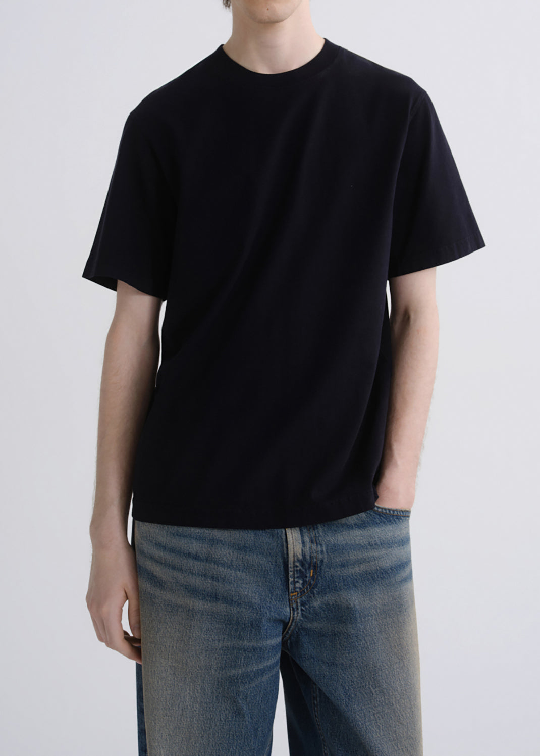 A person wearing a plain black short-sleeve crew-neck T-shirt and blue jeans, standing against a grey background.