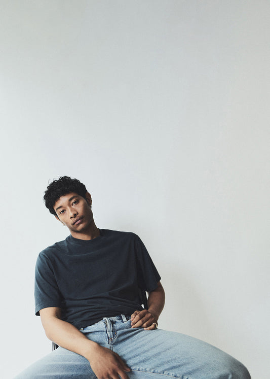 Person wearing a dark t-shirt and light jeans sitting against a plain background