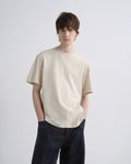 A young man wearing a cream-colored short-sleeve crewneck t-shirt and dark blue jeans, standing against a light gray studio background.