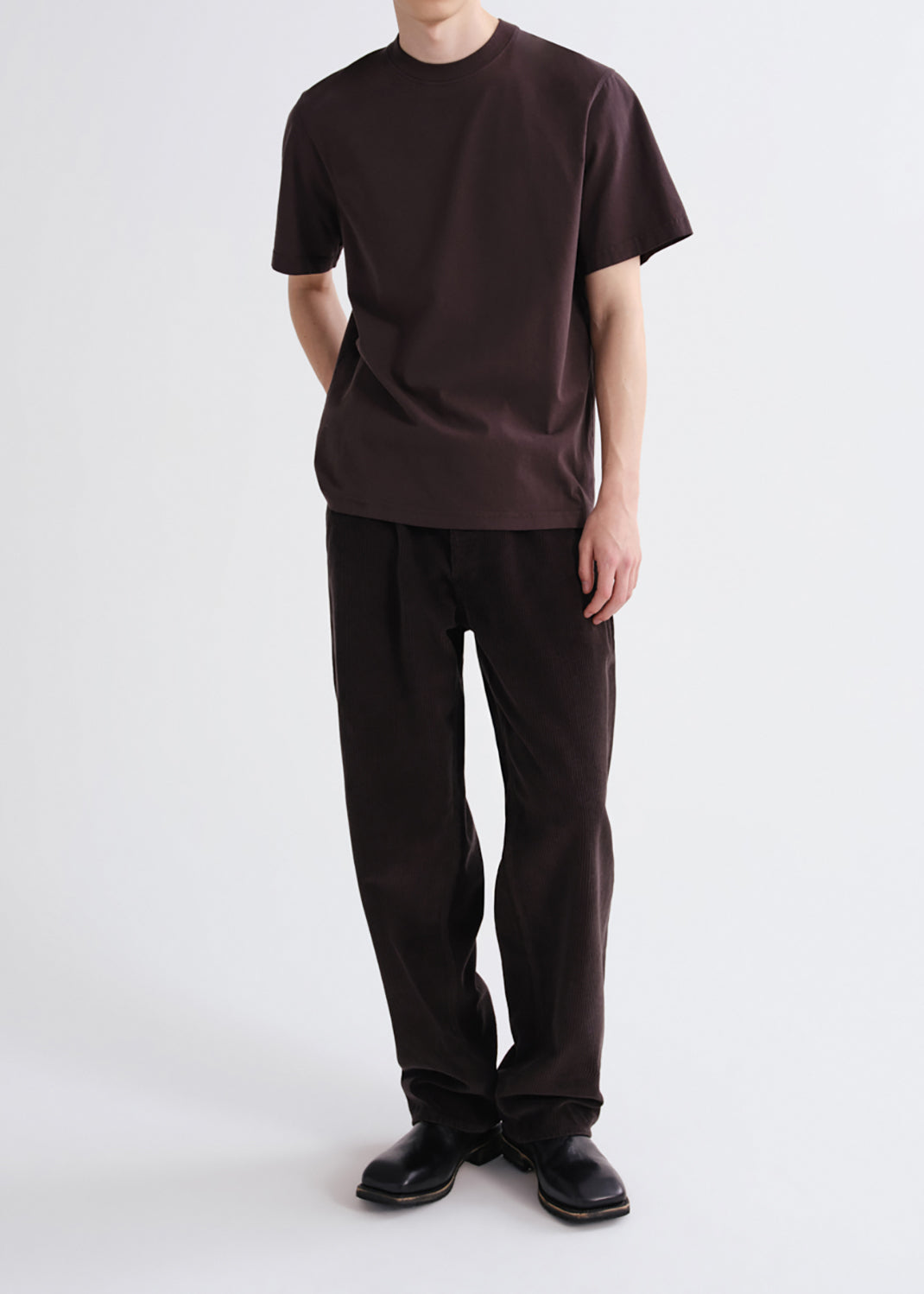 A model wearing a dark brown short-sleeve crewneck t-shirt and matching dark brown relaxed-fit trousers, paired with black leather shoes, against a grey background.