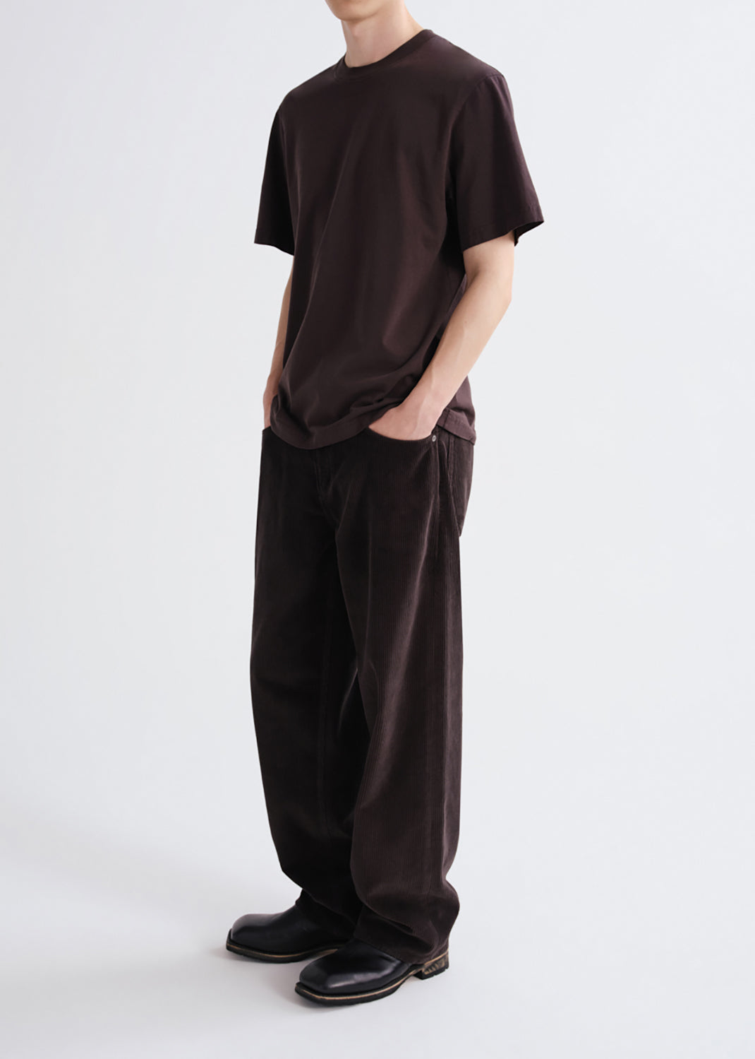 A model wearing a dark brown short-sleeve t-shirt and matching dark brown wide-leg trousers, paired with black leather shoes, photographed against a grey background.