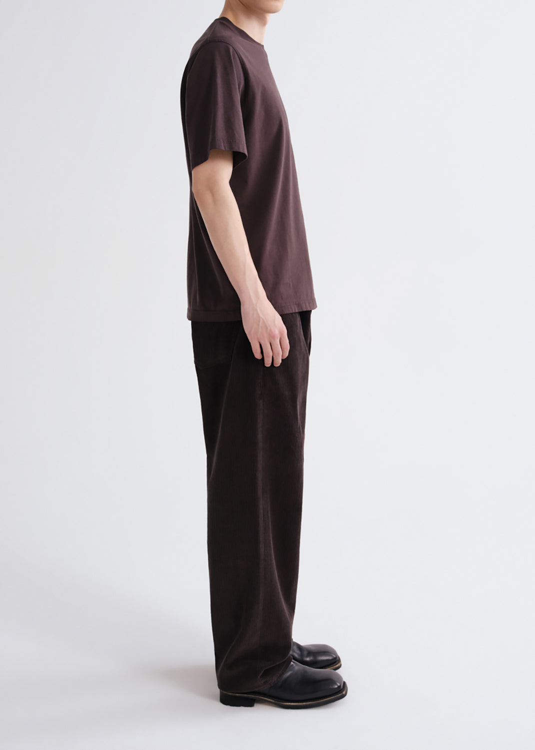A model shown from the side wearing a dark brown short-sleeve t-shirt, matching dark brown wide-leg trousers, and black leather shoes against a grey background.