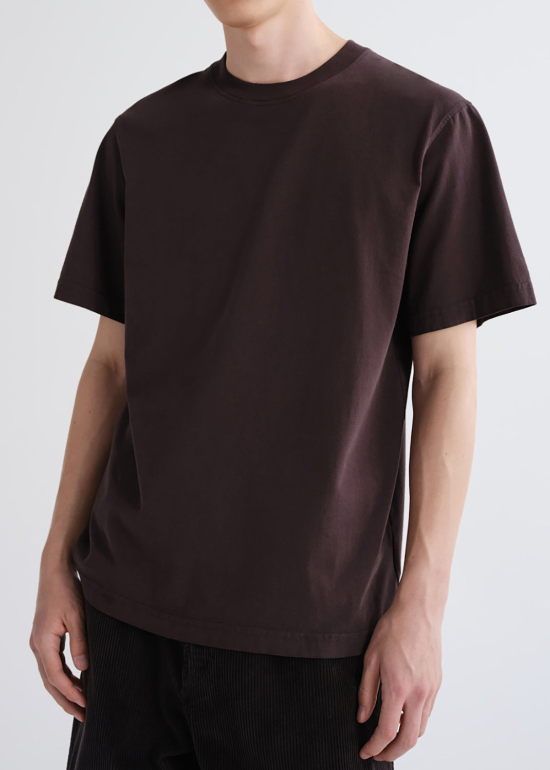 A man wearing a dark brown short-sleeve crewneck t-shirt and black corduroy pants, shown from the neck down against a grey background.
