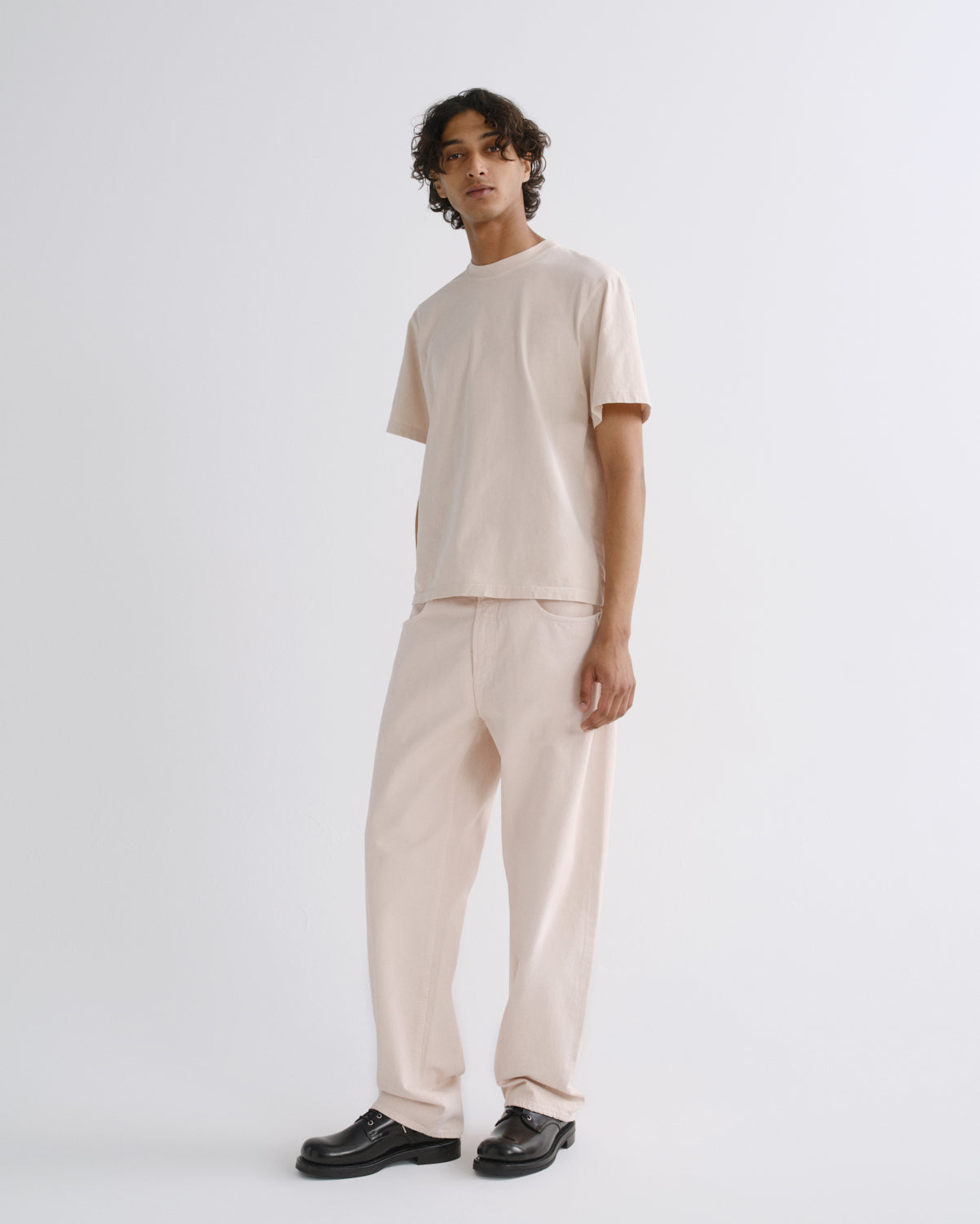 A model stands in a studio wearing a light beige short-sleeve t-shirt and matching wide-leg trousers, paired with black leather shoes, against a plain light gray background.