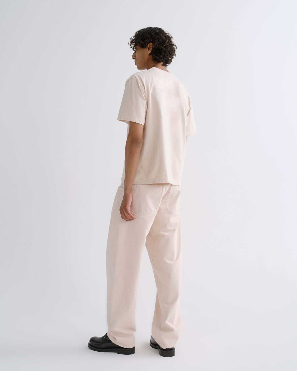 A model is shown from the back wearing a matching light beige short-sleeve t-shirt and wide-leg trousers set, paired with black shoes, against a plain light gray studio background.