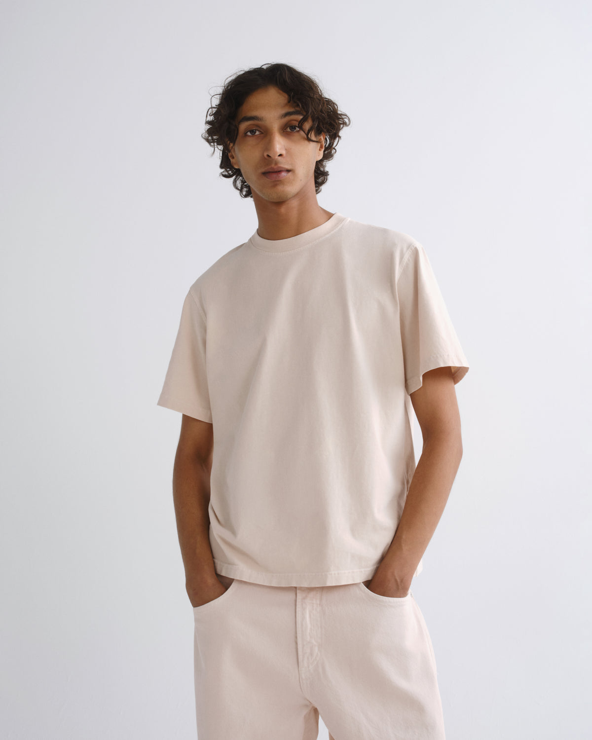 A man wearing a light beige crew-neck t-shirt and matching light beige trousers, standing against a plain light gray studio background.