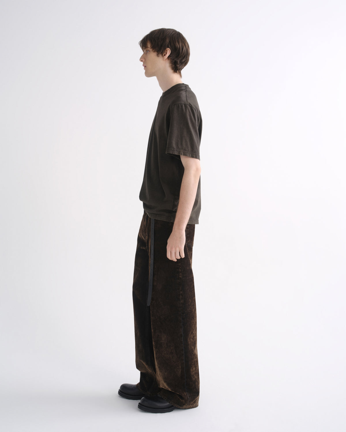 Person wearing a dark brown short sleeve T-shirt with dark brown pants, side view, on a grey background