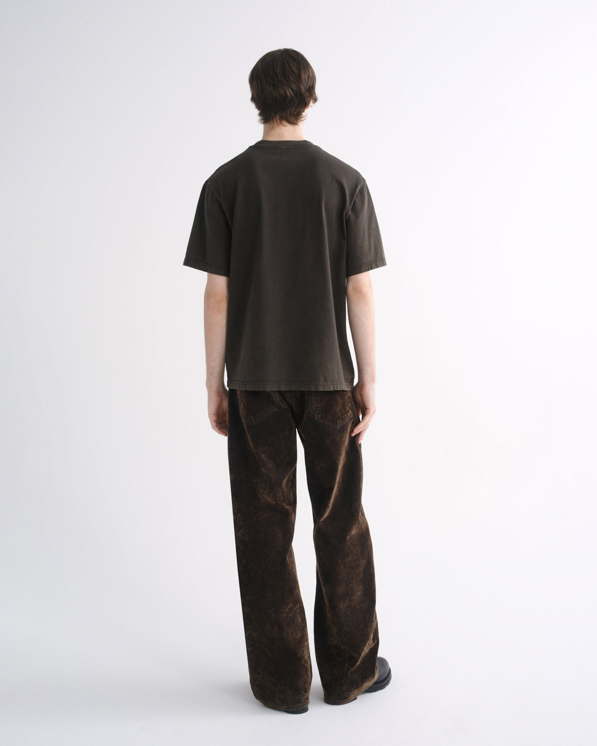 Person wearing a dark brown short sleeve T-shirt with dark brown pants, back view, on a grey background