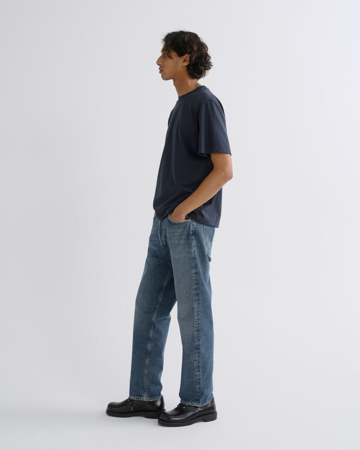 A man standing in profile wearing a dark navy blue t-shirt, blue jeans, and black leather shoes against a light gray studio background.