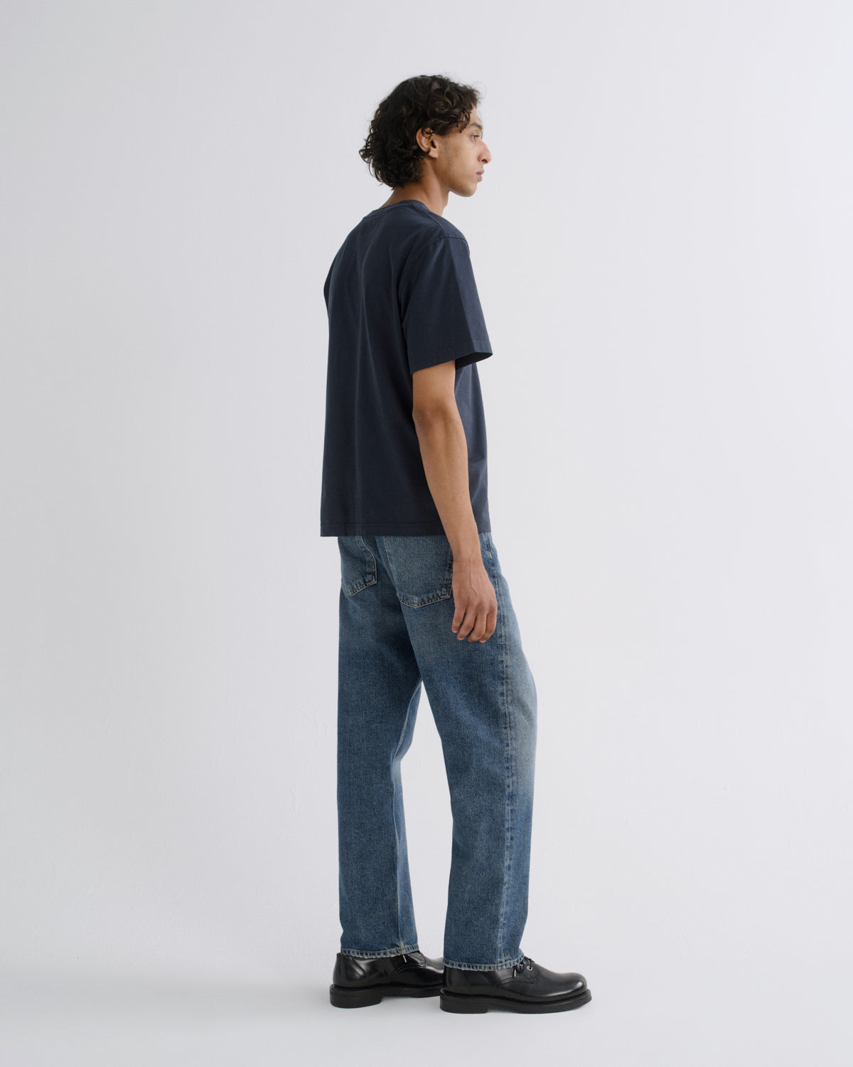A man wearing a dark navy blue t-shirt, blue jeans, and black leather shoes, shown from a side profile against a light gray studio background.