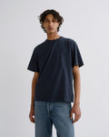A man wearing a dark navy blue crew-neck t-shirt and light blue jeans, standing against a plain light gray studio background.
