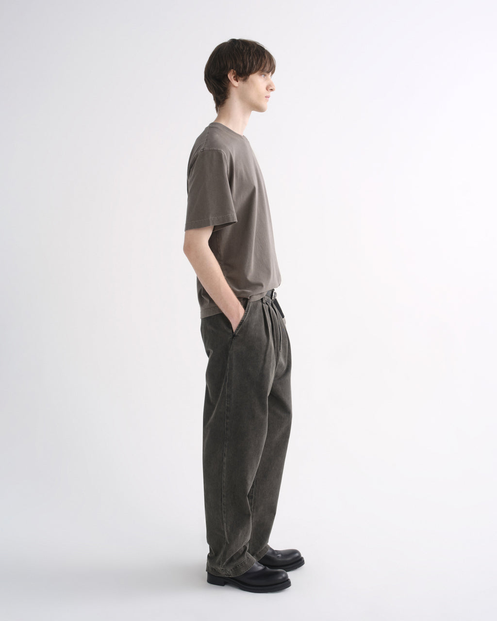 Person wearing a grey short sleeve T-shirt with dark pants and black shoes, side view, on a grey background