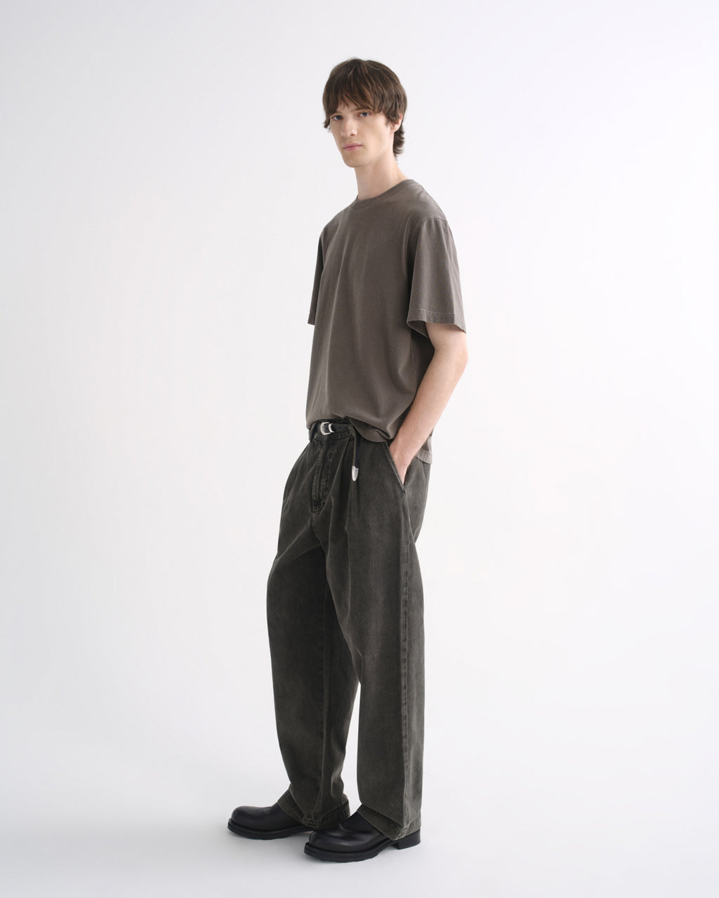 Person wearing a grey short sleeve T-shirt with dark pants and black shoes, side view, on a grey background