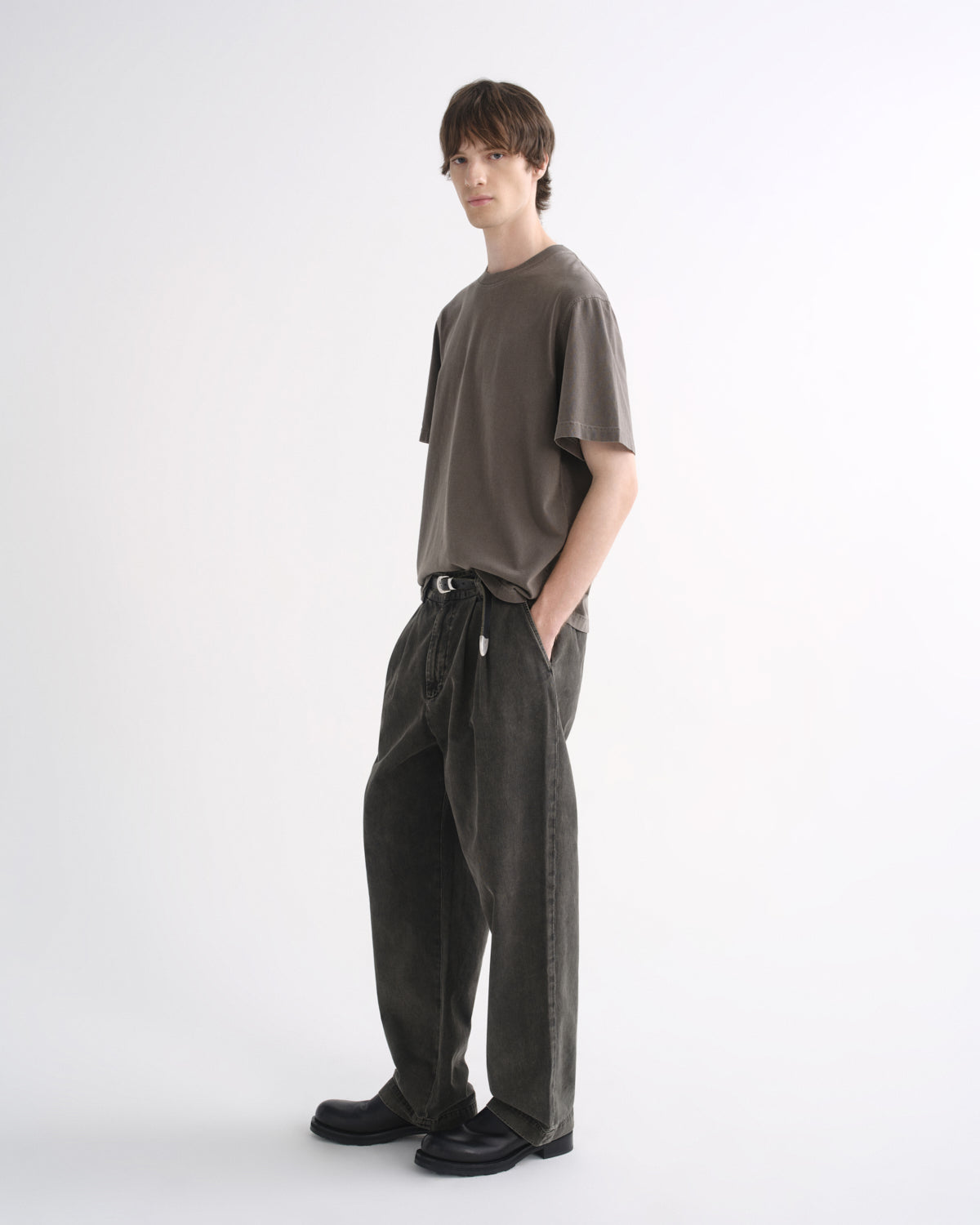 Person wearing a grey short sleeve T-shirt with dark pants and black shoes, side view, on a grey background