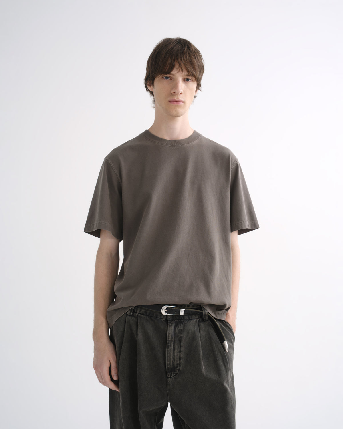 Person wearing a grey short sleeve T-shirt with dark pants and black shoes on a grey background