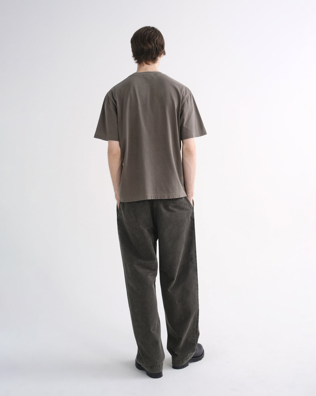 Person wearing a grey short sleeve T-shirt with dark pants and black shoes, back view, on a grey background