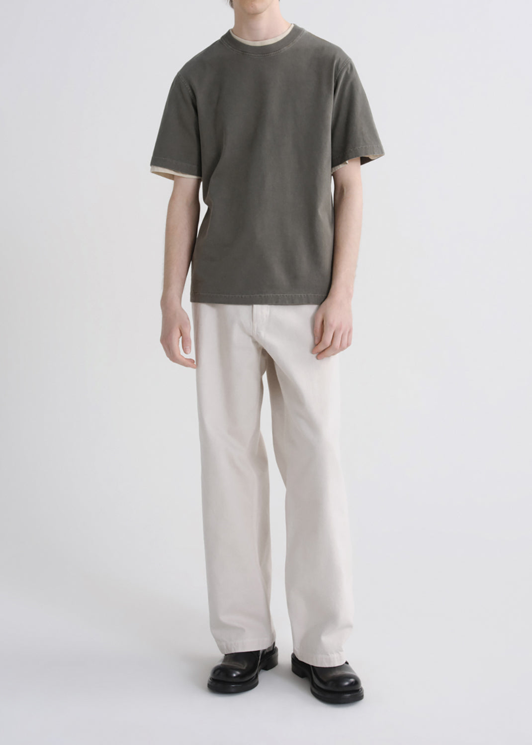 A model wearing a dark olive green short-sleeve t-shirt with cream ribbed trim, paired with off-white straight-leg trousers and black leather shoes, shot against a grey background.