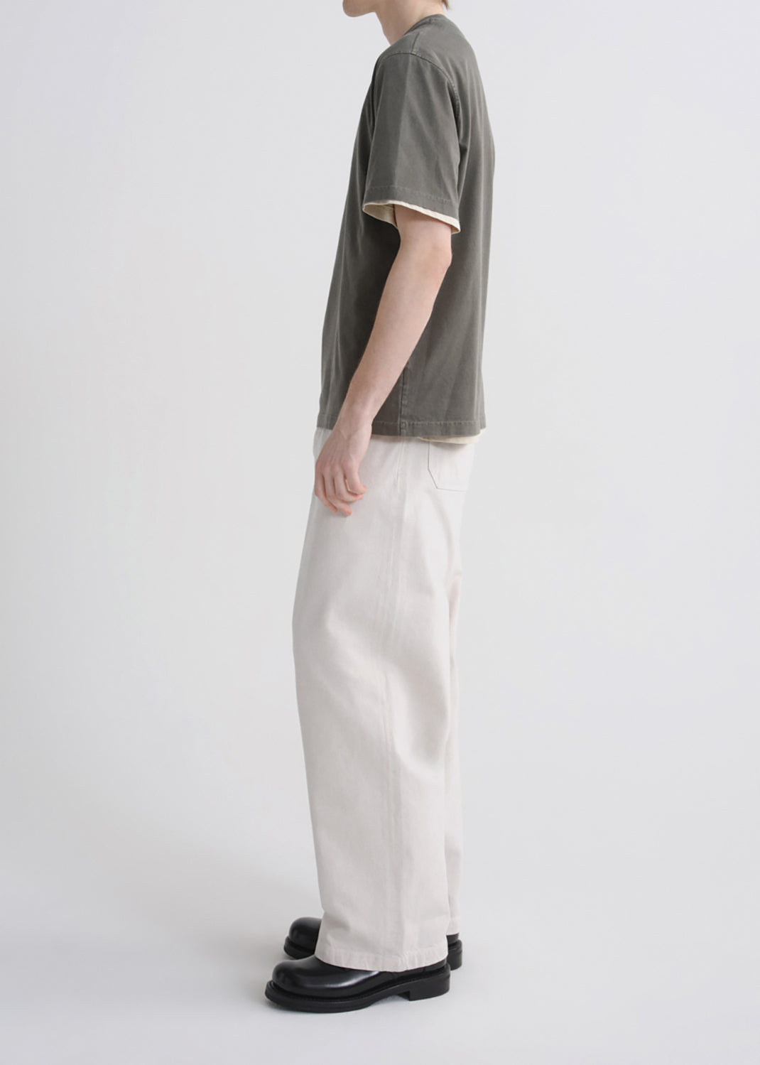 A side-profile studio shot of a model wearing a muted olive green short-sleeve t-shirt with cream ribbed trim, off-white wide-leg trousers, and black leather shoes against a grey background.