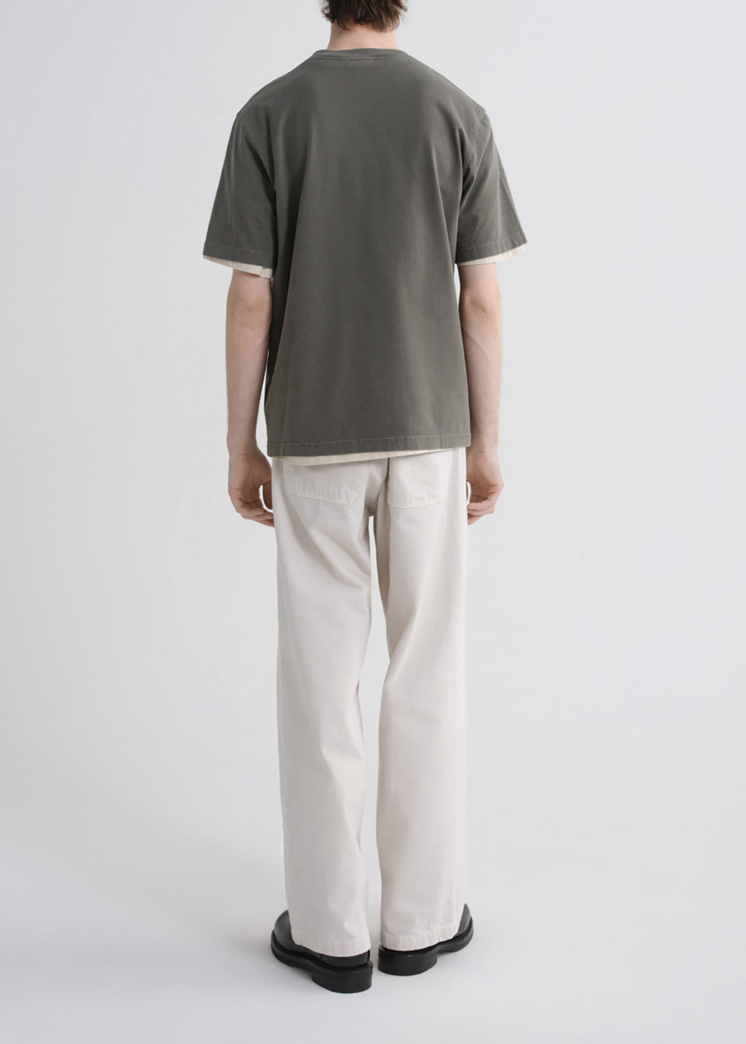 A person shown from the back wearing a muted olive green short-sleeve t-shirt and off-white wide-leg trousers, standing against a grey background.