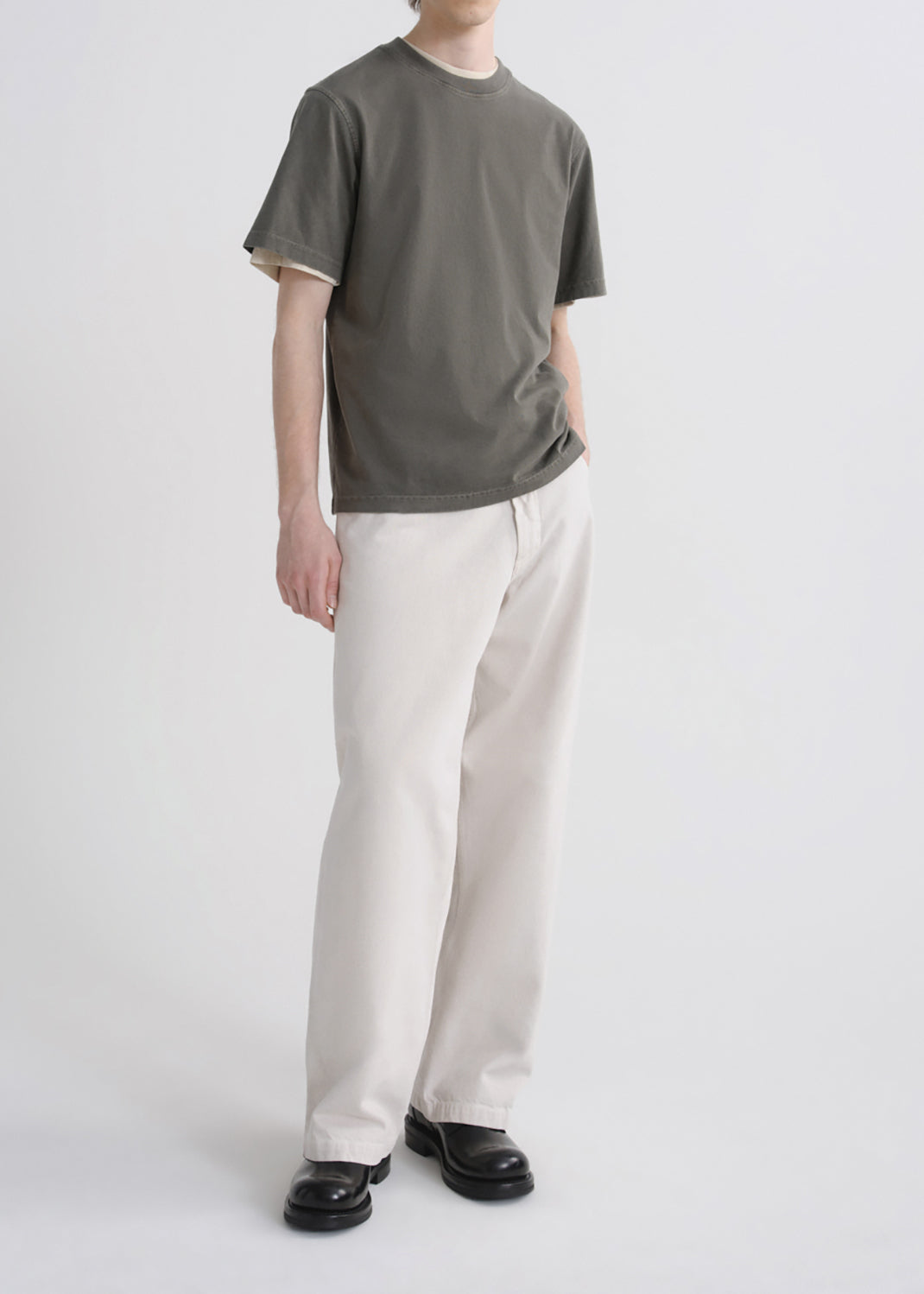 A model wearing a dark olive green t-shirt with contrasting cream ribbed collar and sleeve cuffs, paired with off-white relaxed-fit trousers and black leather loafers, shot against a seamless grey  background.