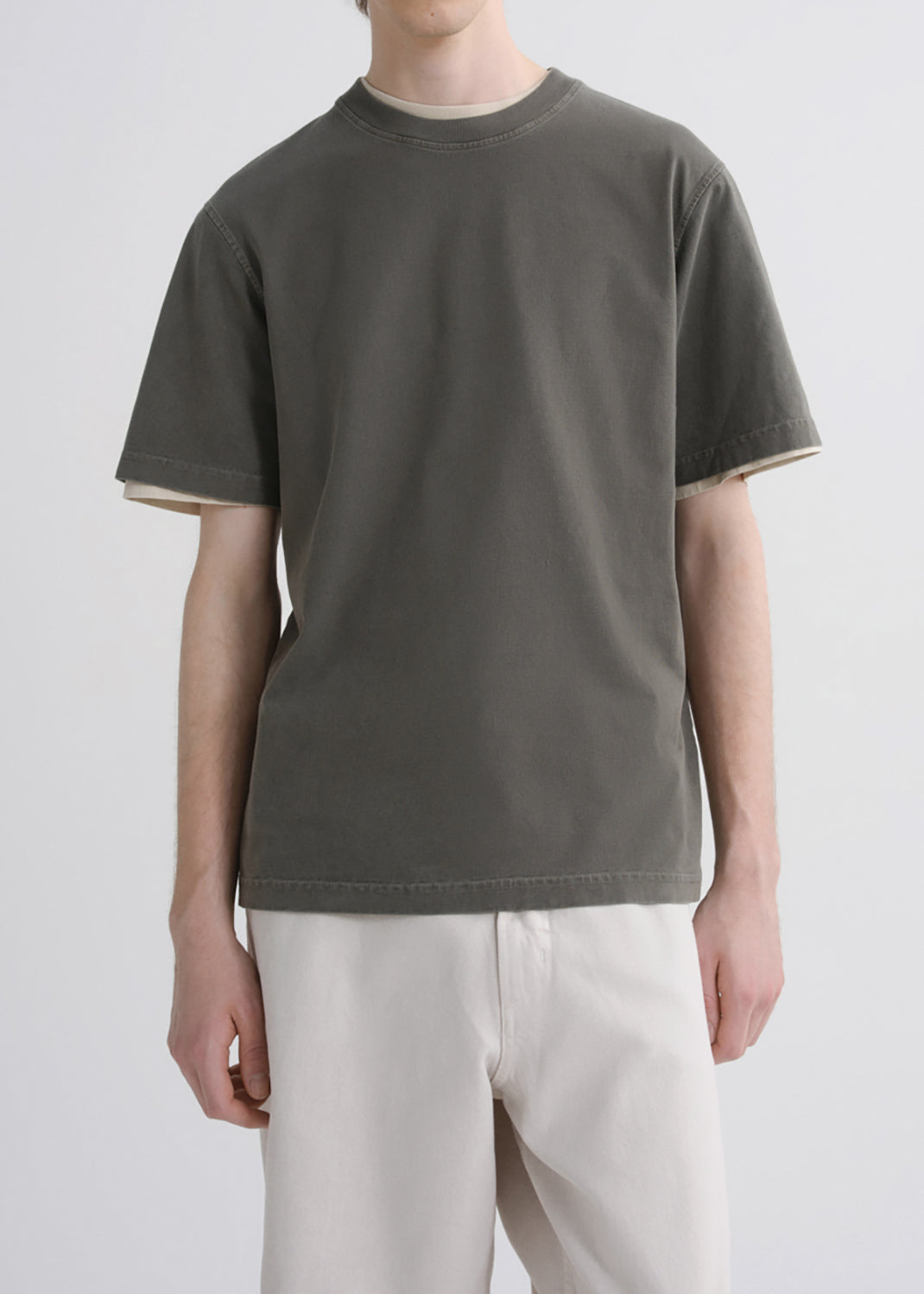 A person wearing an oversized short-sleeve crew-neck t-shirt in dark olive green with light beige contrast trim on the collar and sleeve cuffs, paired with off-white trousers, against a grey background.