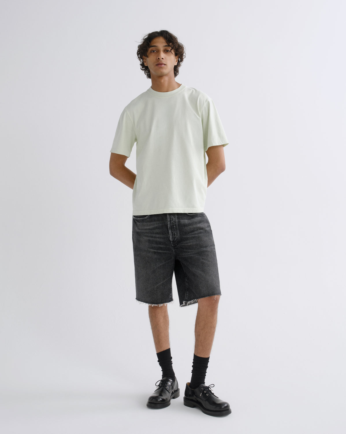 A man modeling a light sage green short-sleeve t-shirt, dark gray denim shorts, black socks, and black leather shoes against a plain light gray studio background.