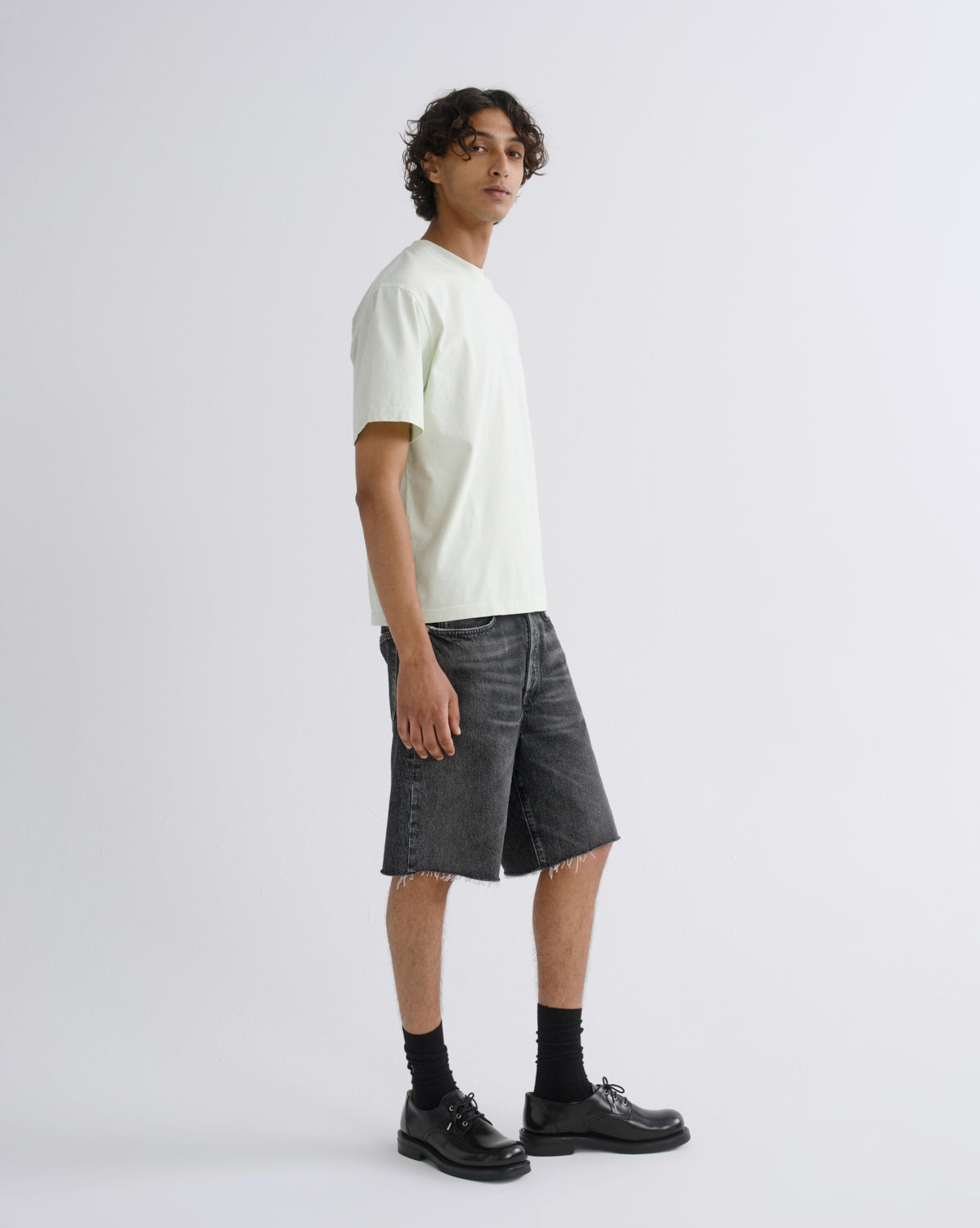 A man stands in profile wearing a pale cream short-sleeve T-shirt, dark gray washed denim shorts, black socks, and black leather shoes against a light gray studio background.