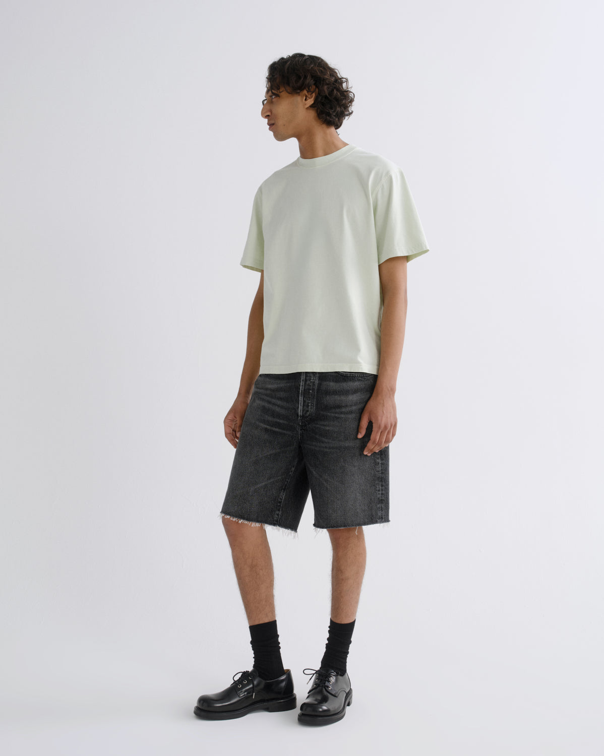 A model stands in profile wearing a pale mint green short-sleeve t-shirt, dark gray washed denim shorts, black socks, and black leather shoes against a light gray studio background.