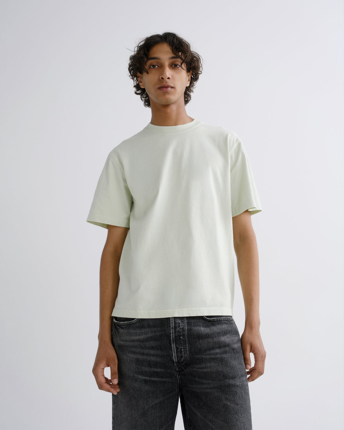 A model wearing a pale sage green crewneck t-shirt and dark gray jeans, standing against a light gray studio background.