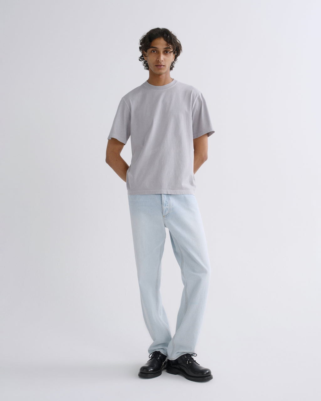 A man modeling a light gray crewneck t-shirt and light blue jeans with black shoes, standing against a plain light gray studio background.