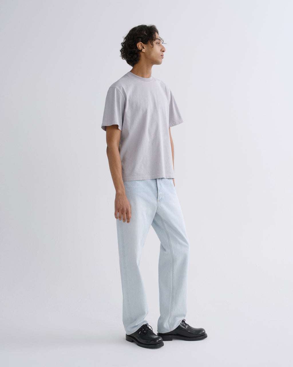 A man wearing a light gray crewneck t-shirt, light blue relaxed-fit jeans, and black leather dress shoes, standing in profile against a solid light gray studio background.