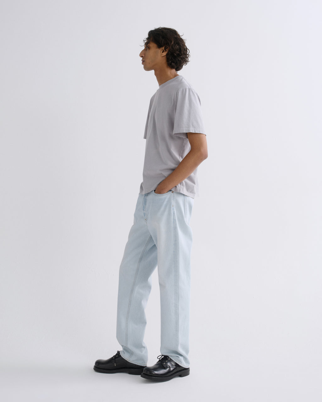 A man in a light gray t-shirt and light blue relaxed-fit trousers, standing in profile against a plain white studio background.