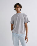 A man wearing a light gray short-sleeve crewneck t-shirt and light blue jeans, standing against a plain light gray studio background.