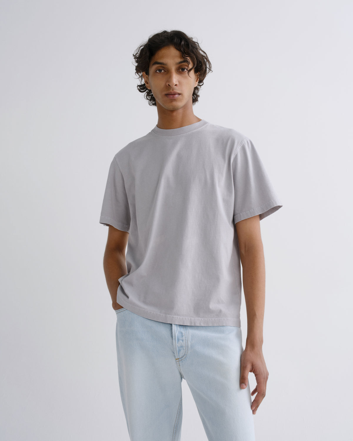A man wearing a light gray short-sleeve crewneck t-shirt and light blue jeans, standing against a plain light gray studio background.
