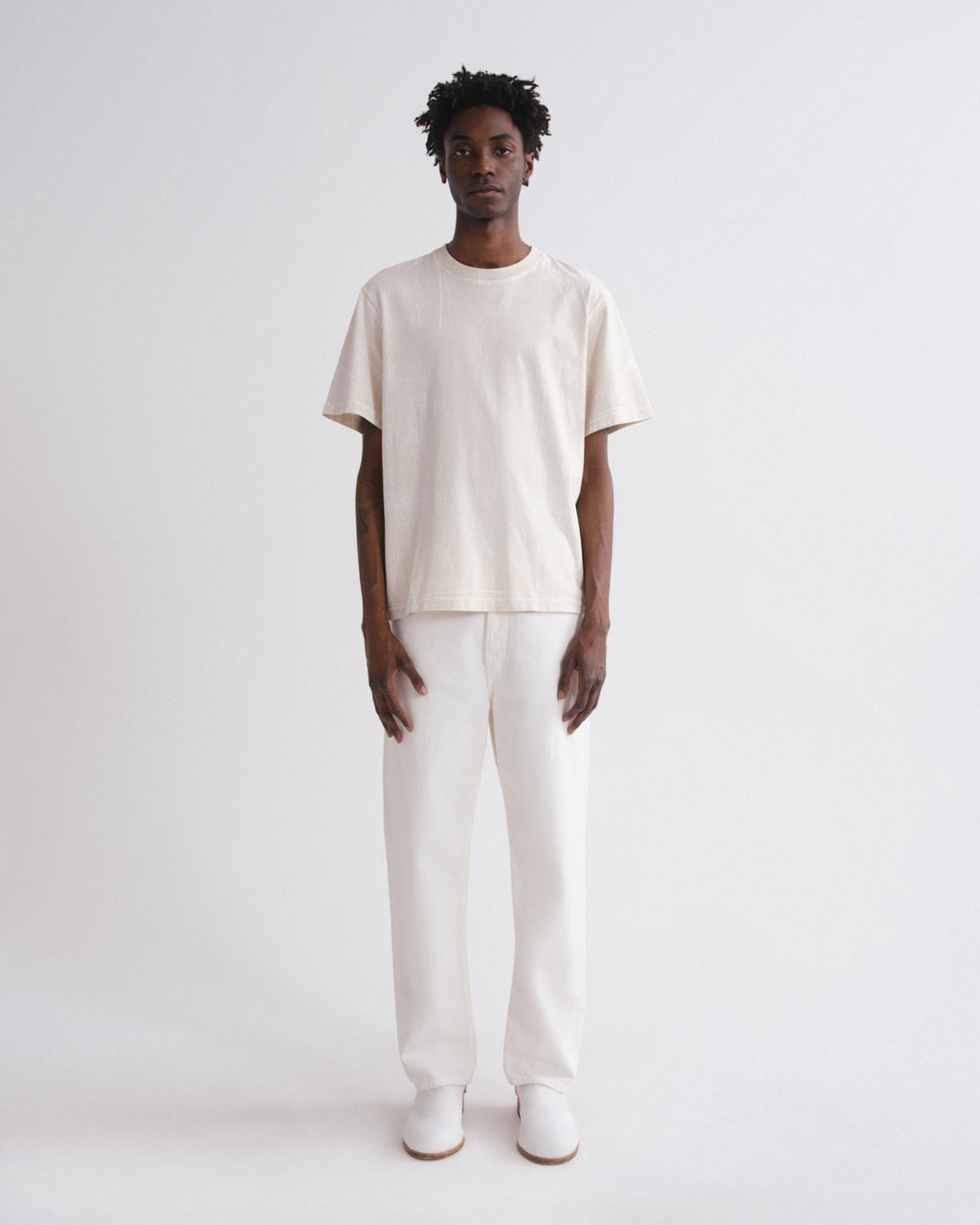 A model stands against a plain light gray studio background wearing a loose off-white short-sleeve t-shirt, matching off-white trousers, and white shoes.