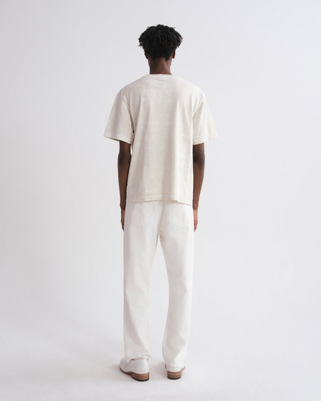 A model viewed from behind wearing a cream-colored short-sleeve t-shirt and matching cream-colored trousers, standing against a plain white studio background.