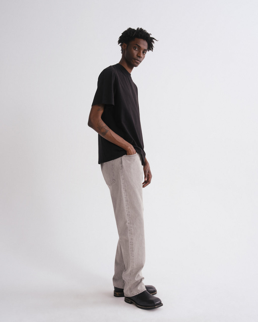 A man stands in profile wearing a black short-sleeve t-shirt, light beige relaxed-fit trousers, and black shoes against a plain white studio background.